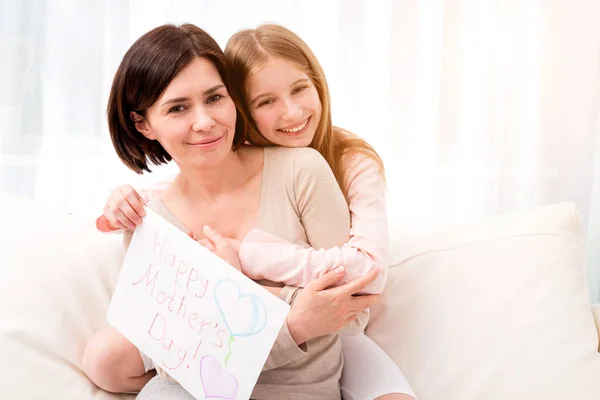 Kleines Mädchen umarmt ihre Mutter und hält eine Glückwunschkarte in der Hand — Stockfoto