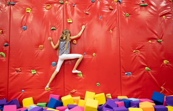 Little smiling blond girl standing near red climbing wall in the play room — Stock Photo, Image