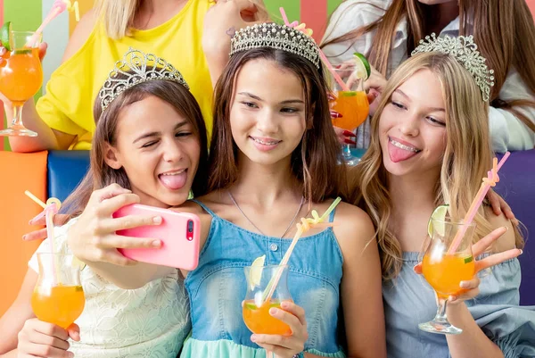 Красивые улыбающиеся девочки-подростки в платьях и коронах сидят, обнимаясь, держа напитки — стоковое фото