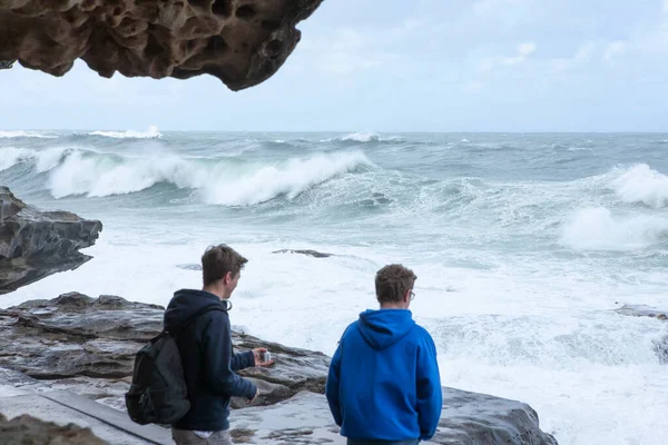 Two guys watching the storm waves crashing on the rocks, Bondi Australia