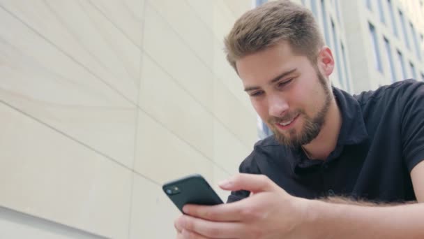 Человек с бородой сидит и пользуется телефоном в городе — стоковое видео