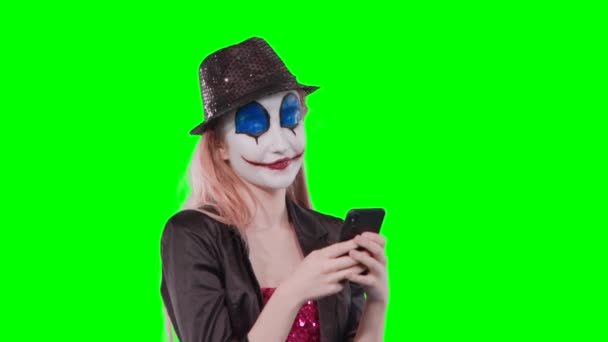 緑の背景に分離された携帯電話を使用してハロウィーン道化メイクで幸せな陽気な金髪女の肖像 — ストック動画