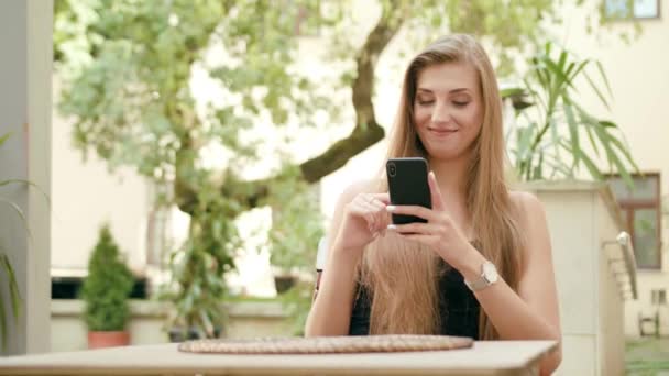 Jovencita usando un teléfono en la ciudad sentada a la mesa — Vídeos de Stock