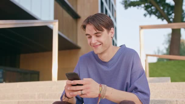 Νεαρός κάθεται και χρησιμοποιώντας ένα τηλέφωνο στην πόλη — Αρχείο Βίντεο