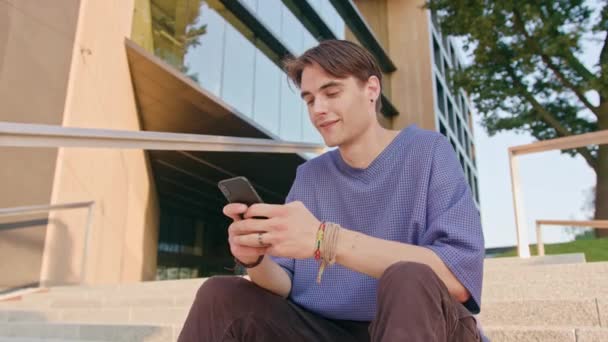 Νεαρός κάθεται και χρησιμοποιώντας ένα τηλέφωνο στην πόλη — Αρχείο Βίντεο
