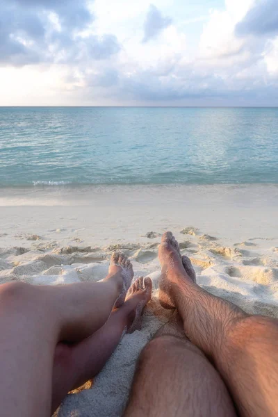 Las piernas de una joven pareja enamorada, que se sientan en la playa y ven el atardecer —  Fotos de Stock