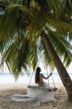 Beyaz elbiseli bir gelin büyük bir palmiye ağacının altında salıncakta sürüyor..