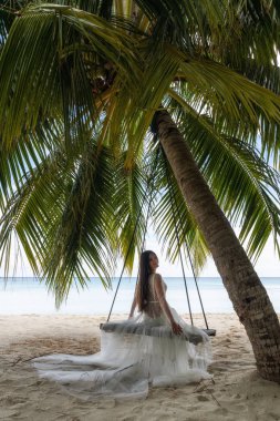 Beyaz elbiseli bir gelin büyük bir palmiye ağacının altında salıncakta sürüyor..