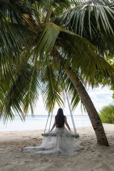 Seorang pengantin dalam gaun putih naik di ayunan di bawah pohon palem besar . Stok Gambar Bebas Royalti