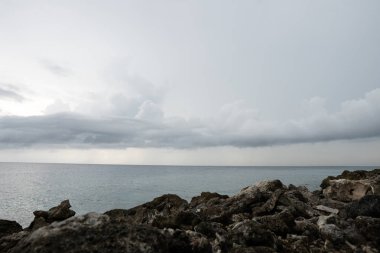 Ön planda kayalık bir kıyı var. Uzakta, okyanus ve bulutlar.
