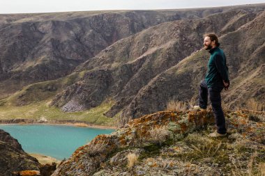 Genç bir adam bir uçurumun kenarında duruyor ve valley adlı bir nehir ile görünüyor. Dağ manzarası. Bahar. Ili Nehri. Kazakistan