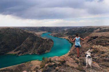 Nehir kenarındaki uçurumda köpeği olan genç bir kız. Nehirli muhteşem bir dağ manzarası. İlkbaharda Ili Nehri. Kazakistan. Almaty Bölgesi