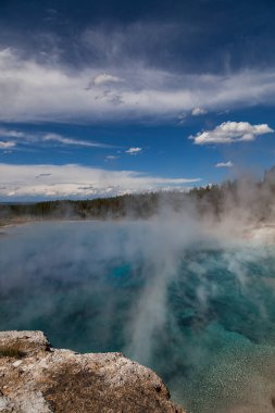 Excelsior Geyser 'in mavi jeotermal suyu beyaz buharla yükseliyor yüksek ağaçların kenarından yükseliyor ve mavi gökyüzüne doğru Yellowstone Milli Parkı' ndaki beyaz bulutlarla birlikte..