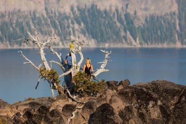 Tanımlanamayan iki kadın, öğleden sonra güneşinde krater gölüne bakan kayalık bir tepede büyüyen çoğunlukla ölü bir çam ağacının üzerinde duruyor..