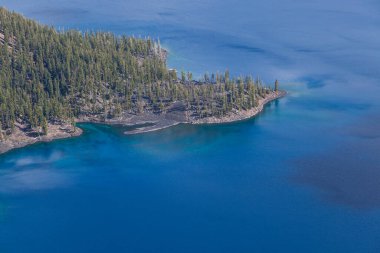 Büyücü Adası 'nın batı kıyısına bakan volkanik bir kaya yüzeyi, Crater Gölü Ulusal Parkı' nda mavi ve turkuaz sularda büyük yeşil ağaçlar serpiştiriyor..
