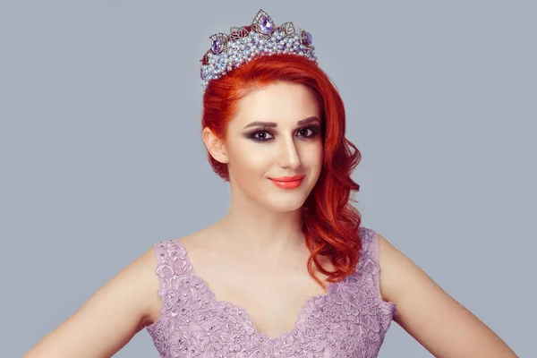 Confident beauty pageant queen. Serious redhead woman in crown with purple pearls and crystals in violet color lace dress vogue style with curly wavy hair curls looking at you camera purple background