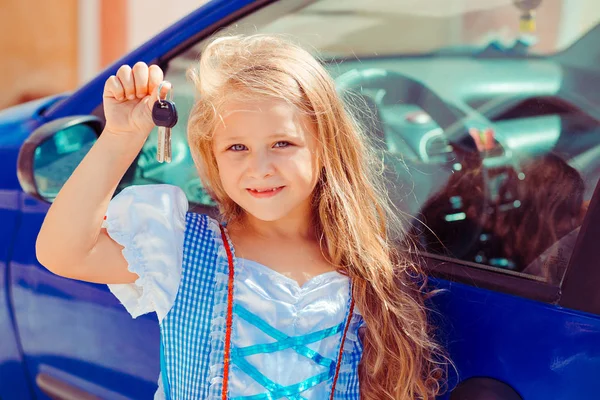 Pretty girl with long golden hair wearing princess dress and showing keys standing near new blue car in sunlight