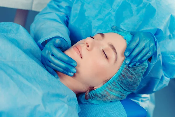 Skin face and care concept, Doctor hands medical beautician makes facial massage holds patient face examination before cosmetic plastic surgery procedures in a clinic surgery room; O.R background.