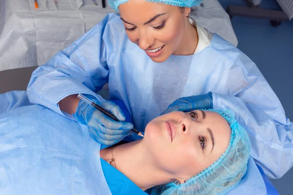 Anti-aging treatment and face lift. Beautiful doctor woman is about to draw lift lines on patient neck with pencil for lifting procedure. Two people in a surgery clinic room, patient lying down