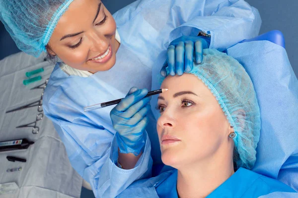 Anti-aging treatment face lift. Beautiful doctor woman about to draw lift line on patient forehead with pencil for anti-wrinkles lifting procedure. Two people in surgery clinic room patient lying down