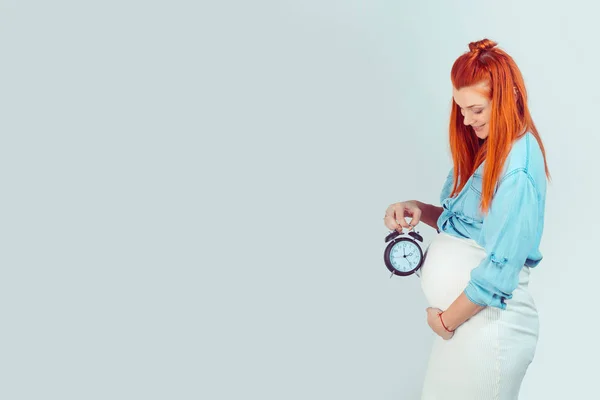 Clock is ticking, expectation concept. Side view of smiling young woman with baby in tummy holding alarm clock waiting for birth and smiling at camera on blue. Mixed race model, hispanic irish woman