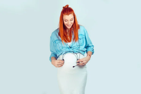 Happy redhead girl holding headset and letting baby in tummy to listen to music smiling looking at belly isolated on light blue background. Mixed race model, latin hispanic irish woman