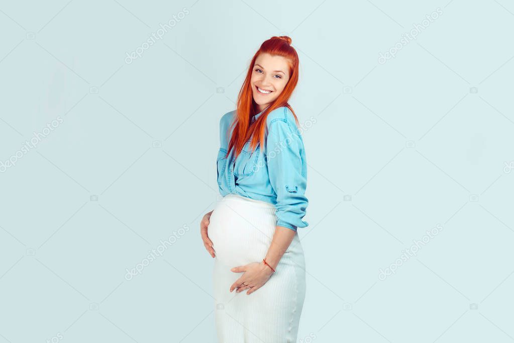 Happiness is inside me. Adorable pretty woman with redhead hairstyle embracing pregnant belly laughing happy while standing on light blue background and giving smile at camera.