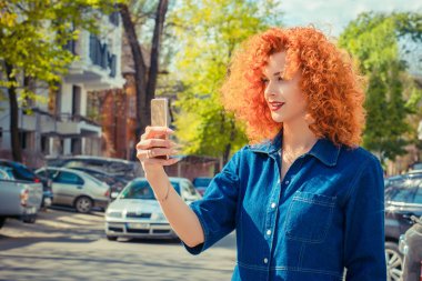 Kendi kendine portre. Bir smartphone kullanarak ve gülümseyen güzel kadın up için kapsama arka planda yeşil ağaçlar şehir dışında seyir telefon ile selfie yapma genç güzellik Kızıl saçlı kız