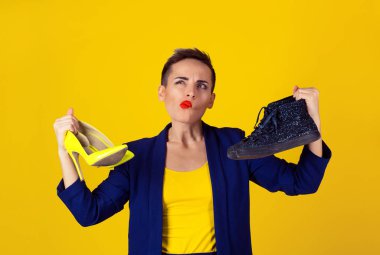 Hangi çifti seçin veya giymek için bilmiyorum. Yüksek topuklu bir çift ve bir düz ayakkabı şaşkın ifade ile yüz tutan kadın üzerinde sarı arka plan izole. Sarı gömlek ve mavi takım elbise modeli.