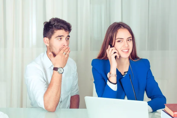 Young shocked man covering mouth while eavesdropping secretly listening juicy gossip conversation of young woman having smartphone call while sitting near at table in the office at workplace or home