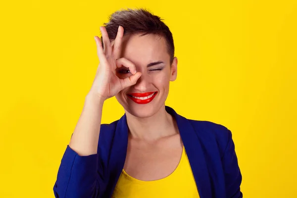 Woman with her fingers over one eye, ok sign hand gesture isolated on yellow background. Model in yellow shirt and blue suit. Satanic 666, Illuminati symbol and sign of the mark of the beast on eye.