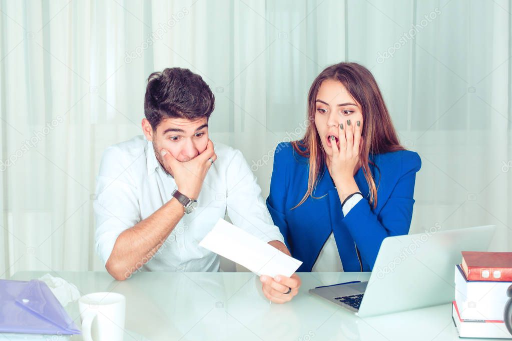 Young amazed surprised stunned couple, colleagues man and woman watching paper notification with huge shock sitting at table in office, living room at home. Huge bills, tax statement concept.