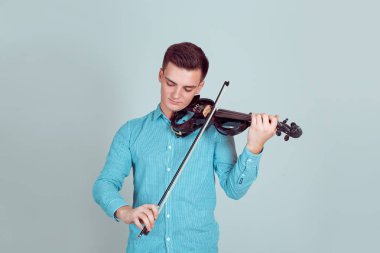 Müzik tutkusu, hobi kavramı. Giyinmiş genç adam modern ve zarif bir stüdyoda ahşap keman oynamaya izole bir ışık mavi duvar arka plan üzerinde. Mavi klasik yatay atış modeli düğmeli gömlek