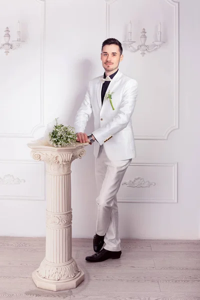Trendy groom in white suit and black shoes standing with bridal bouquet on small column near looking at camera