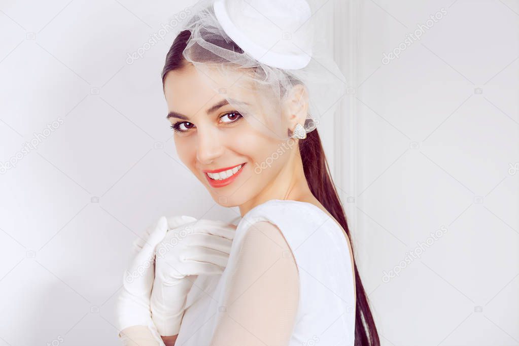 Beautiful smiling brunette in hat with short veil and white dress with gloves smiling charmingly at camera