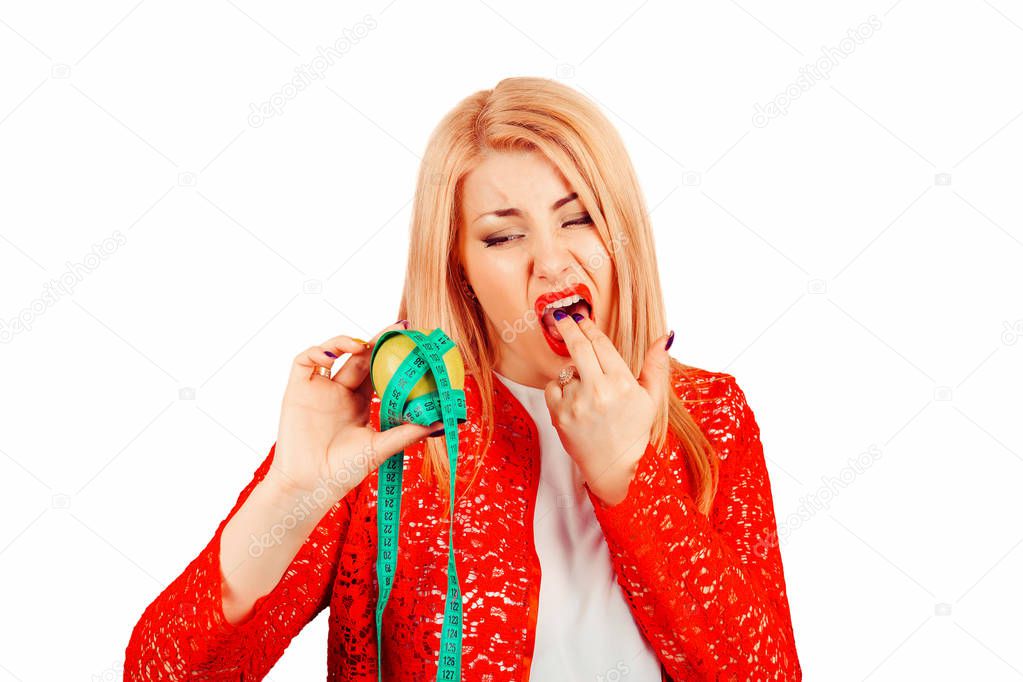 Young woman putting fingers in mouth about to vomit holding measuring tape around apple, annoyed frustrated and fed up sticking her finger in her throat showing she is about to throw up on white background