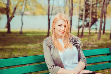 Düşünceli. Şehir Parkı yeşil ağaçlar arka planda bir bankta oturmuş kamera bakarak ciddi bir kadın portresi. Gri ceket beyaz bluz güzel orta yaşlı bayan. Yatay atış ile boşaltmak