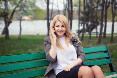 Telefon konuşması. Genç kadın bir şehir parkı yeşil ağaçlar arka planda bir bankta oturmuş cep telefonunda söz. Gri ceket beyaz bluz güzel orta yaşlı bayan. Yatay atış ile boşaltmak