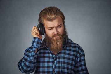 Bir genç ciddi İspanyol DJ Kulaklık dinleme giyen portresi. Mavi ekose kareli gömlek gri arka plan üzerinde Isolated sakallı hippi adam. Negatif yüz ifadesi, insani duyguları.