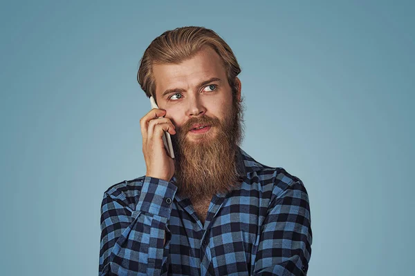 Cool serious man with beard talking on phone and looking up to side. Casual young man talking on smartphone. Hipster male with beard in blue plaid checkered shirt  Isolated on blue studio Background.