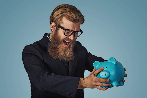 Unhappy man in glasses angry at his piggy bank trying to broke it up, Bearded hipster businessman Isolated on blue Background. Negative face expression, human emotion, body language, reaction attitude