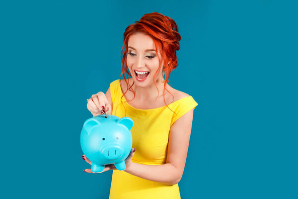 Взволнованная эйфория, когда женщина вставляет монету в копилку, радуется этому. Концепция экономии. Белый человек в жёлтом платье, рыжеволосый, изолированный на синем фоне студии. Экспрессивное лицо
