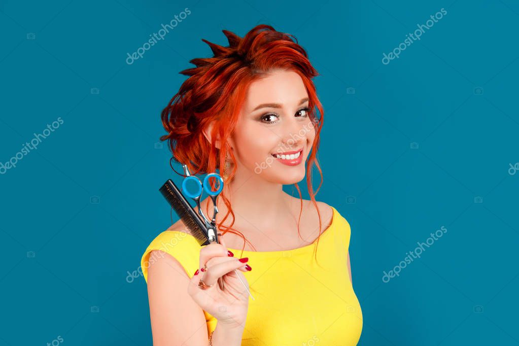 Happy woman holding showing giving scissors and comb. you have to be a hairdresser. Caucasian person in yellow dress redhead hair up isolated on blue studio background. Expressive girl, human emotion