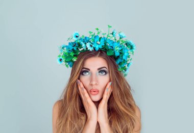 Heyecanlı görüntüsünü açık mavi renkli izole ayakta baş çiçek kafa bandı üzerinde çiçekler olan kadın hayran. Ararken yanaklarda çiçek tacı eller. Şok şok bahar yaz saç modeli