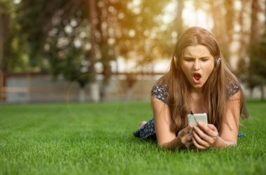 Kötü haber veya sinirli iğrenç duygu ile fotoğraf açık havada yüzünü görmeye telefon bakarak endişeli genç kız baş kulaklıktan yeşil çim üzerinde yatıyor. Çok kültürlü modeli, karışık yarış