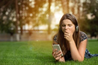 Esneme kadın genç kız açık havada baş kulaklıktan yeşil çim üzerinde yalan bir cep telefonu tutan satır içerik üzerinde sıkıldınız. Çok kültürlü modeli, karışık ırk, Asyalı Rus kız. Kopya alanı.