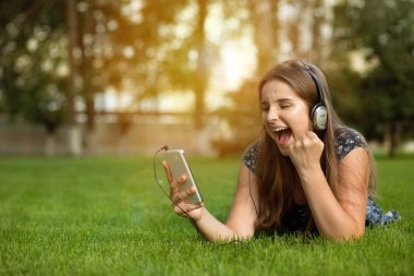 Sevimli genç akıllı telefon elinde tutan sevinçle çığlık atıyor ve mutluluk, sürpriz kazanan ifade ile yüzü açık havada baş kulaklıktan yeşil çim üzerinde yalan. Çok kültürlü modeli, karışık yarış