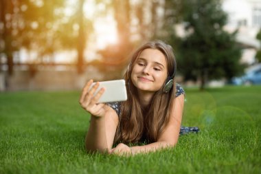 Mutlu genç kız görüntülü sohbet bir selfie poz yapmak bir video mesajı gönderme akıllı telefon seyir tarassut a film, açık havada baş kulaklıktan yeşil çim üzerinde yalan hücre üzerine bir ders.