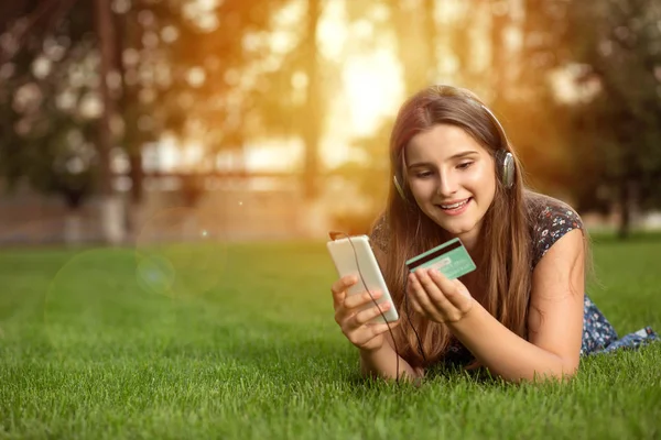 Açık havada baş kulaklıktan yeşil çim üzerinde yalan bir akıllı telefon ile online satın alma mutlu bir kadın portresi. Çok kültürlü modeli, karışık ırk, Asyalı Rus kız. Uzay kopyalayın. İnsan ve teknoloji.