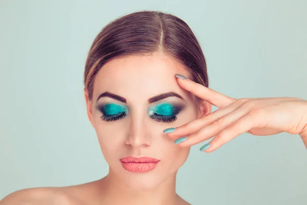 Beautiful woman beauty with artistic blue green eye shadows makeup, holding hand fingers near face eye showing manicure, blue grey nail polish isolated on green background looking down eyes closed